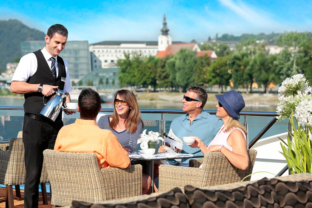 PRIMA_BreakfastTopDeck Waiter serving drinks to guests dining on riverside terrace overlooking church and cityscape