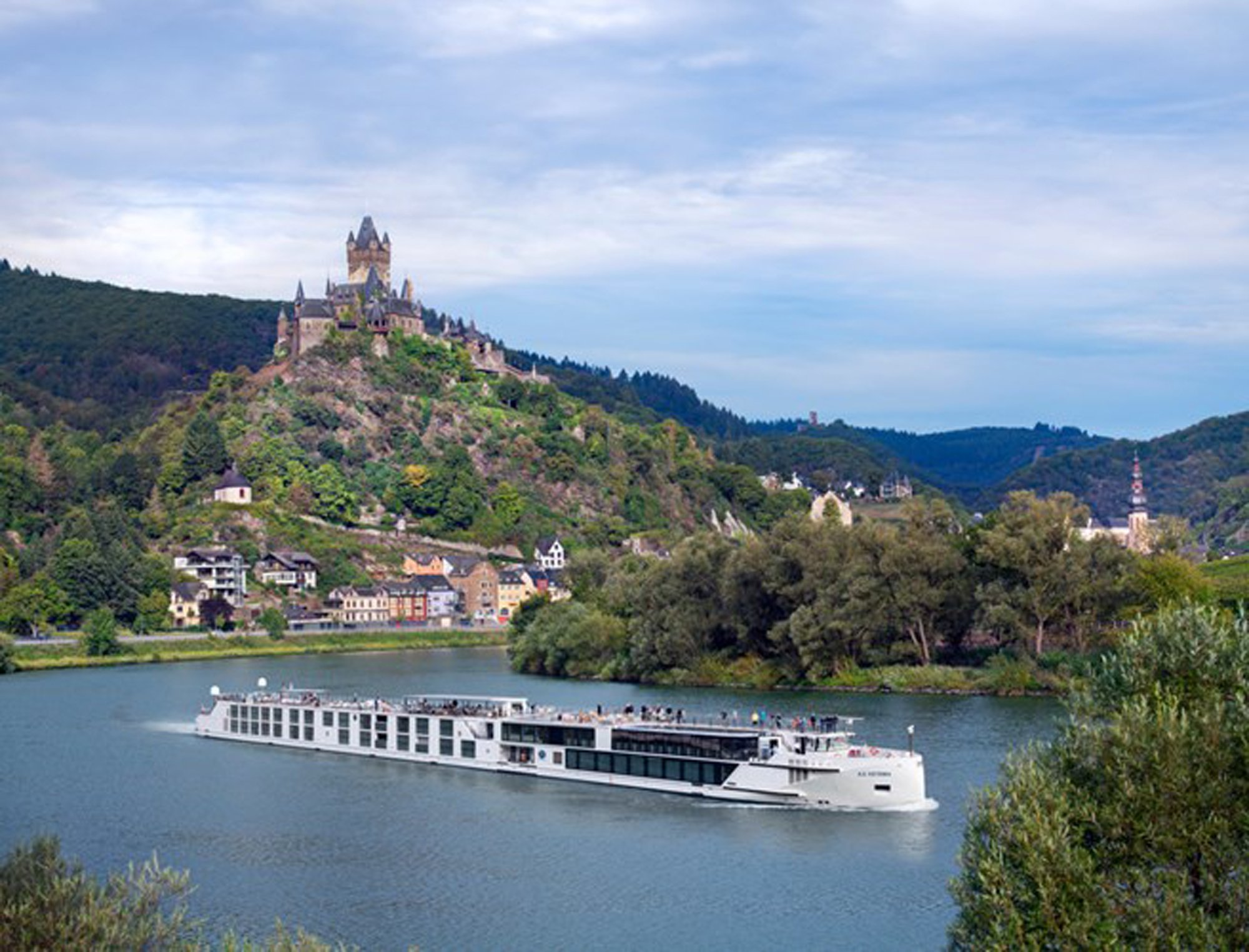 A modern river cruise ship navigates the Moselle River with Reichsburg Castle on the hilltop above Cochem, Germany