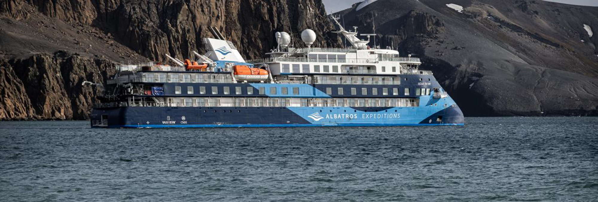 Polar Latitudes expedition cruise ship anchored near a rugged, rocky coastline with steep mountainous terrain
