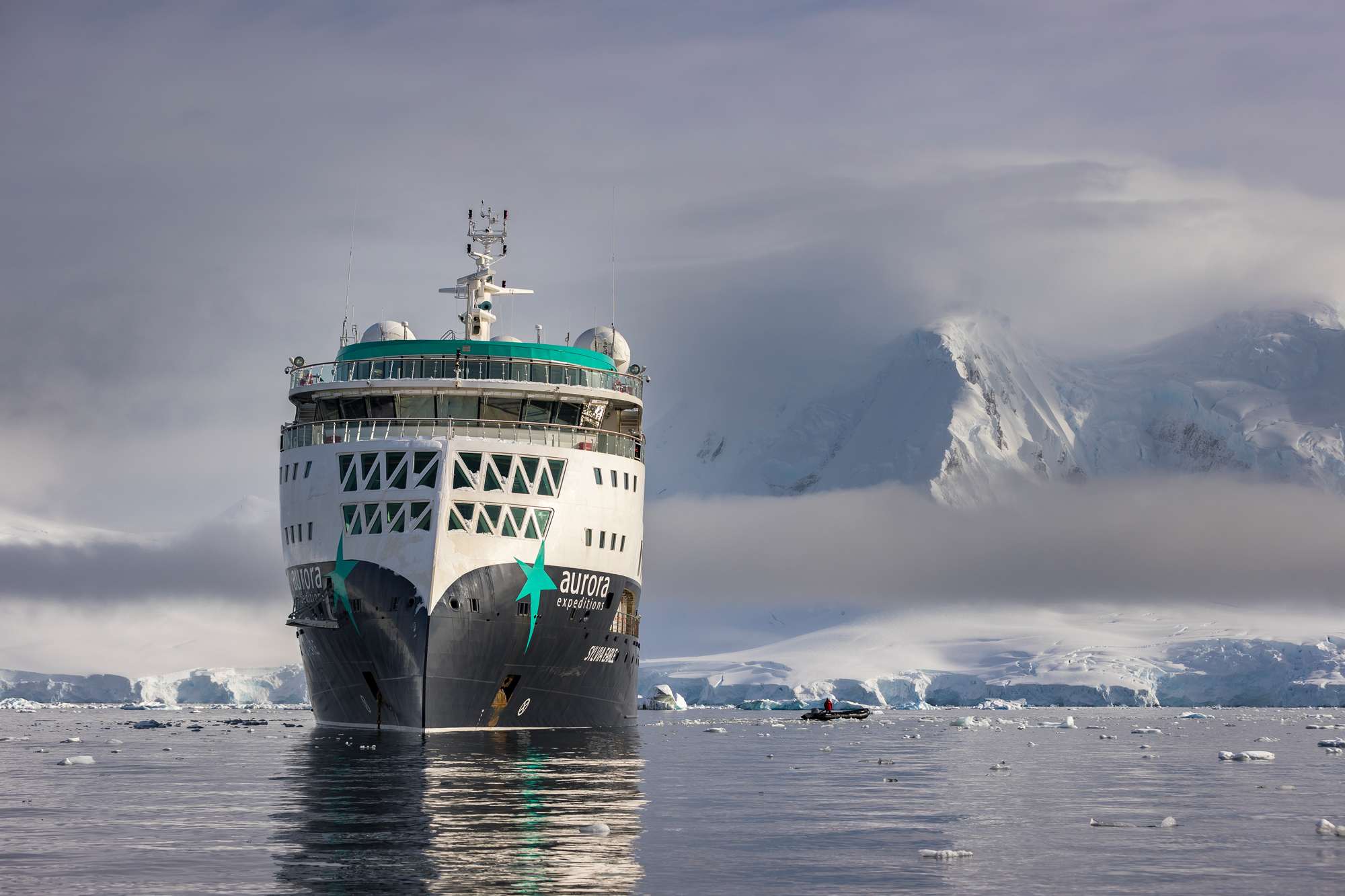 Aurora Expedition Cruise in Antarctic Waters A white and teal expedition cruise ship navigating through icy Antarctic waters, with snow-covered mountains and glaciers in the background and floating ice fragments around the vessel.