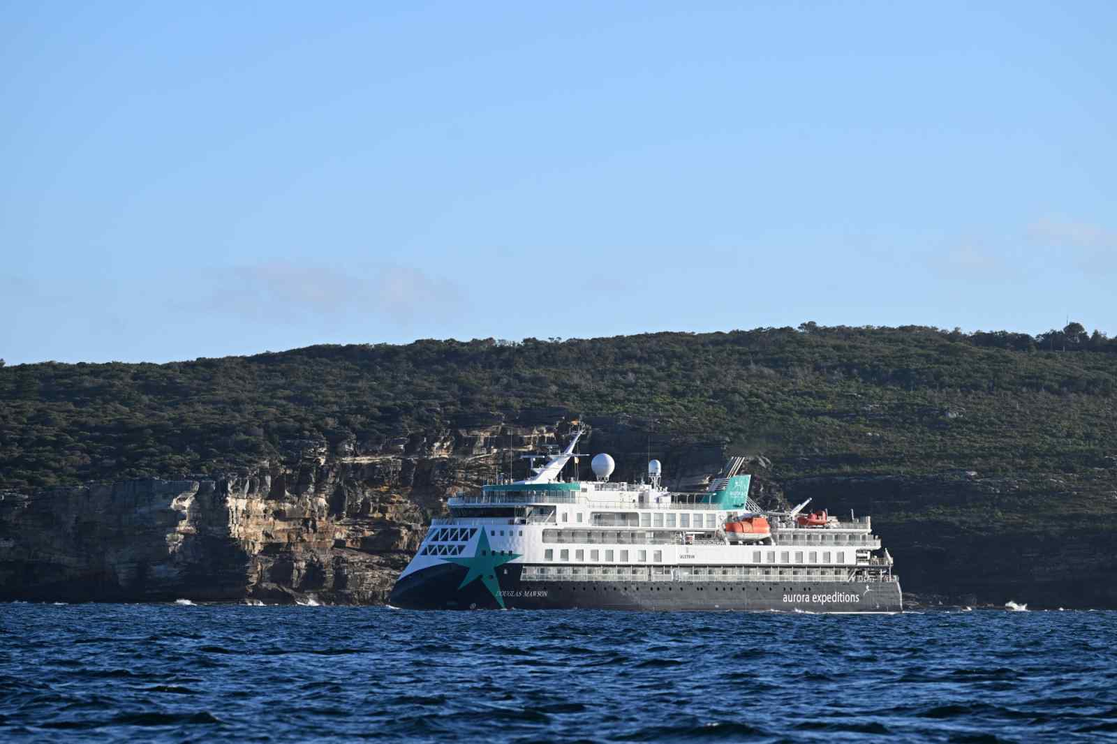 Aurora Expeditions Cruise Ship near Rocky Coast An Aurora Expeditions expedition cruise ship sailing close to a rugged, rocky coastline with dense