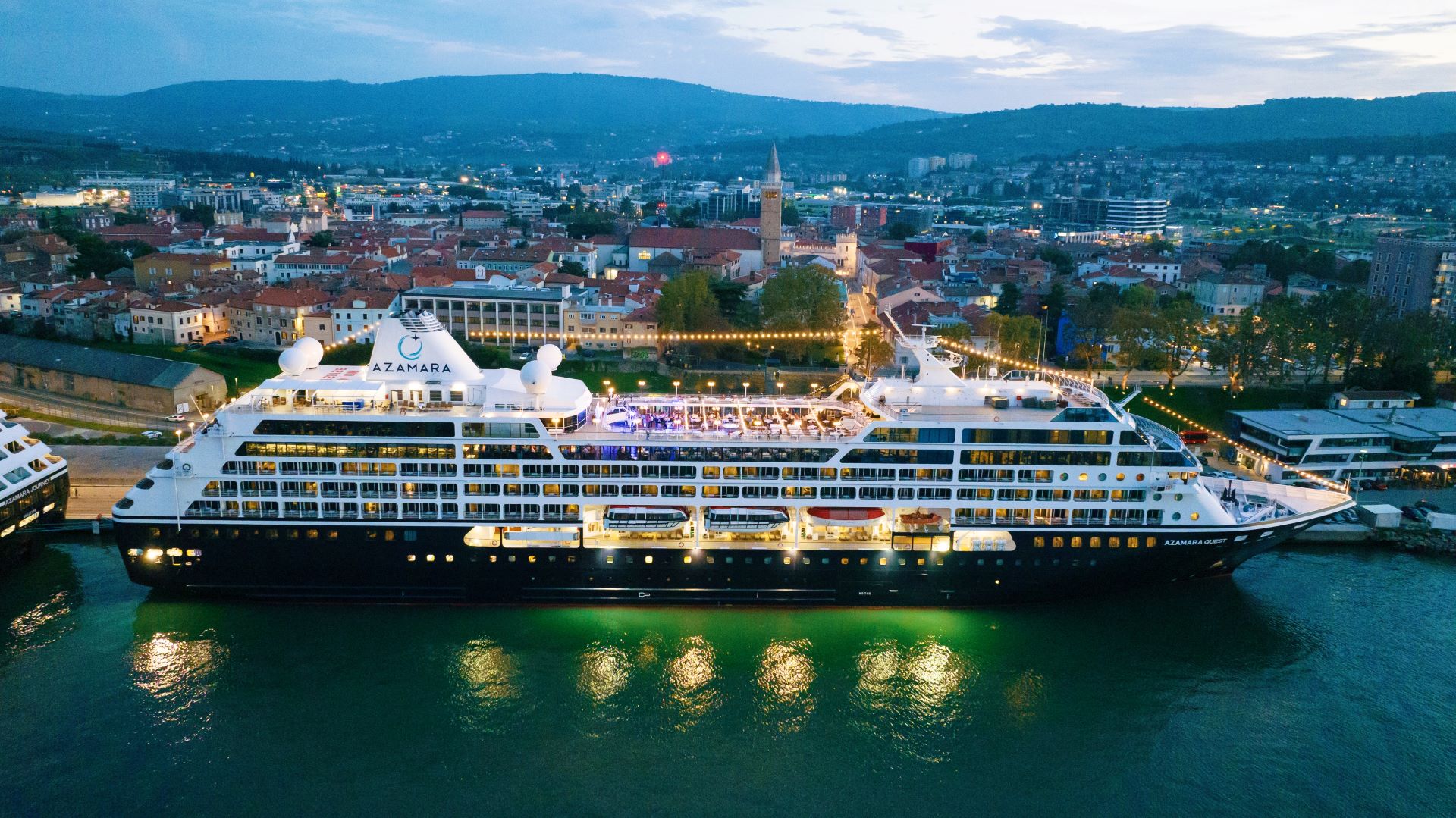 Azamara Cruise Ship Docked in Coastal European City An Azamara cruise ship illuminated at night, docked in a picturesque European city with mountains and urban landscape in the background.
