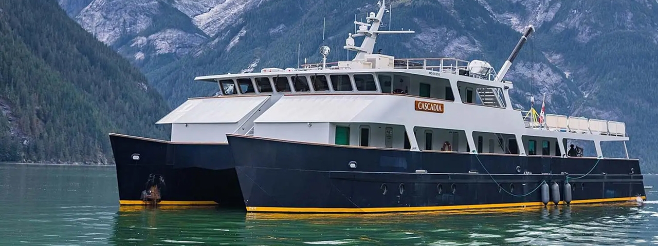 The expedition cruise vessel Cascadia anchored in a pristine glacial fjord surrounded by snow-capped mountains and dense evergreen forests