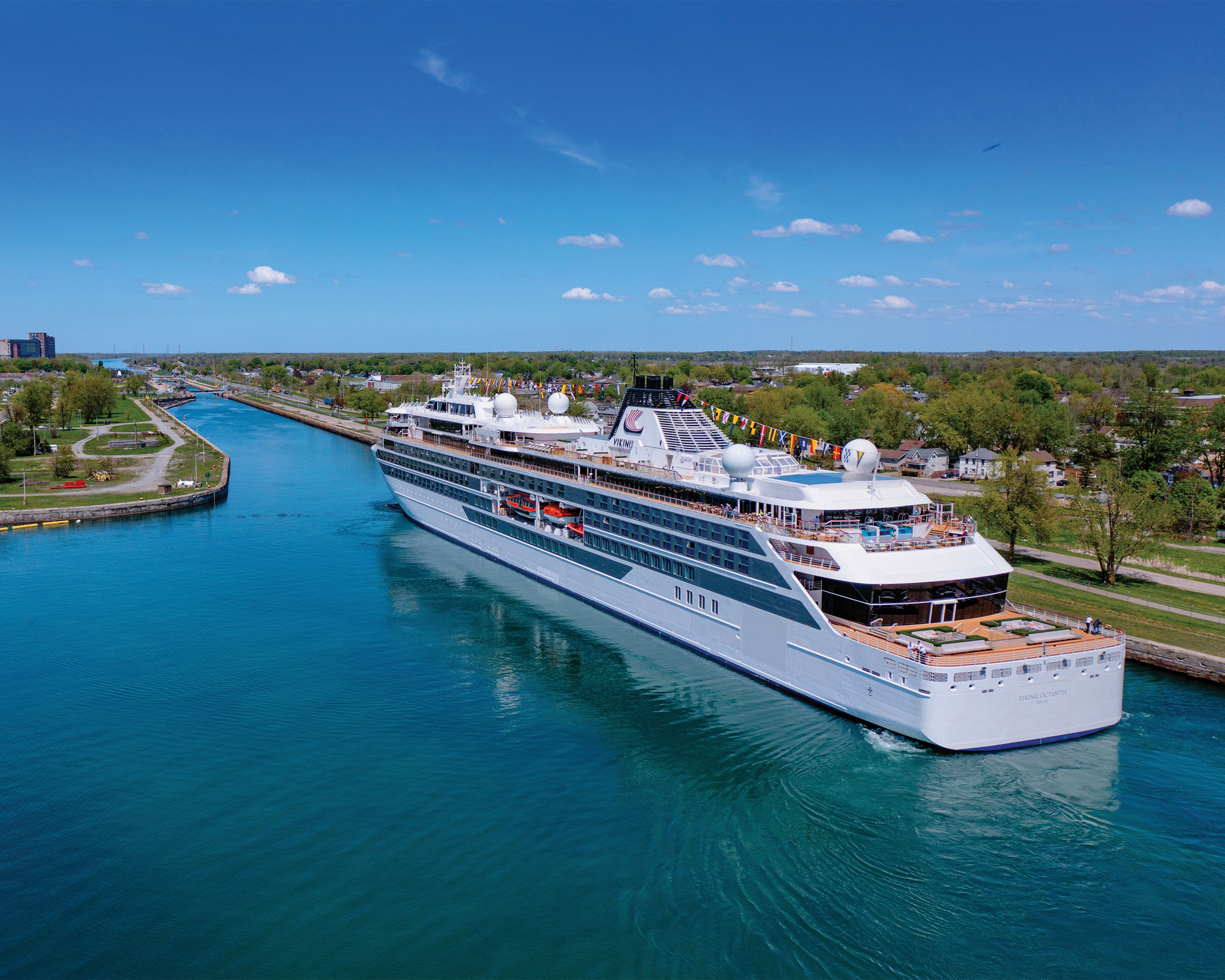 Cruise Ship Navigating Scenic Waterway A sleek white cruise ship sailing through a bright blue canal with lush green shoreline and urban