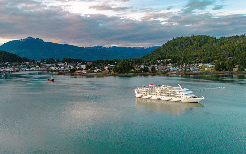 A white cruise ship anchored in a tranquil bay near a forested coastal town with mountains in the background