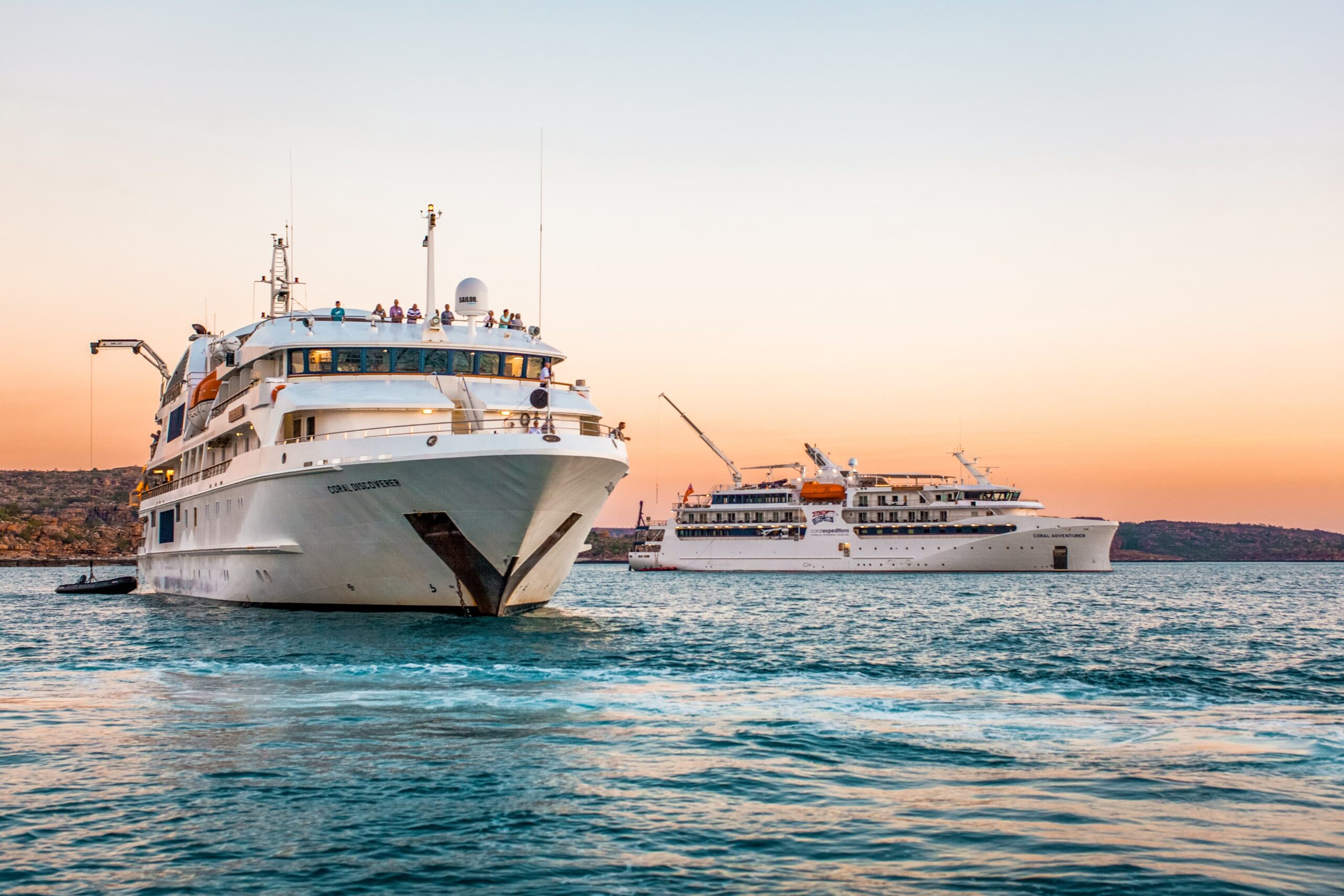 Two white luxury cruise ships anchored in tranquil blue waters during a golden sunset, with rocky