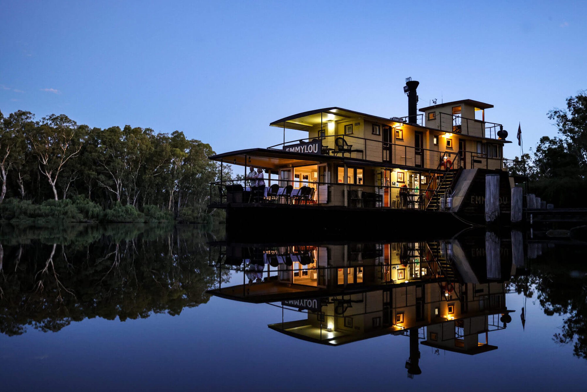 Emmylou Riverboat at Twilight A beautifully illuminated multi-level houseboat named Emmylou glows warmly against the twilight sky, its reflection perfectly mirrored in the still, dark waters below. Surrounded by eucalyptus trees along a serene riverbank, the vessel showcases modern architecture with multiple decks, large windows, and outdoor living spaces.