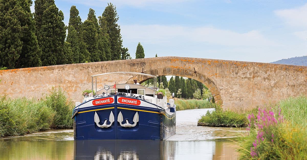 Enchante Barge Passing Under Stone Bridge The MS Enchante, a luxury river cruise barge, navigates through a picturesque European canal