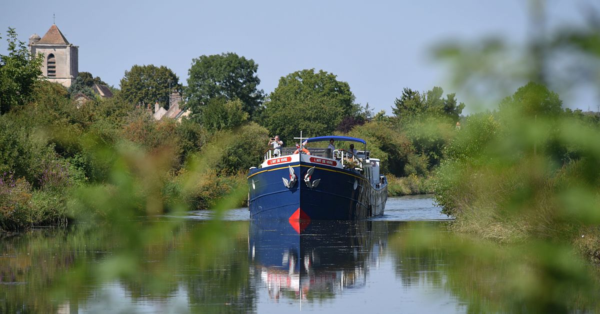 European River Cruise Barge on Waterway A blue and black river cruise barge navigates a peaceful inland waterway lined with lush green vegetation, with a charming church featuring a distinctive bell tower visible in the background among trees.