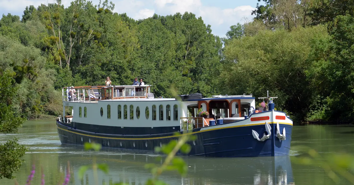 European River Cruise Boat on Waterway A charming European river cruise barge with distinctive blue and white hull navigates a calm waterway surrounded by lush green trees and vegetation.