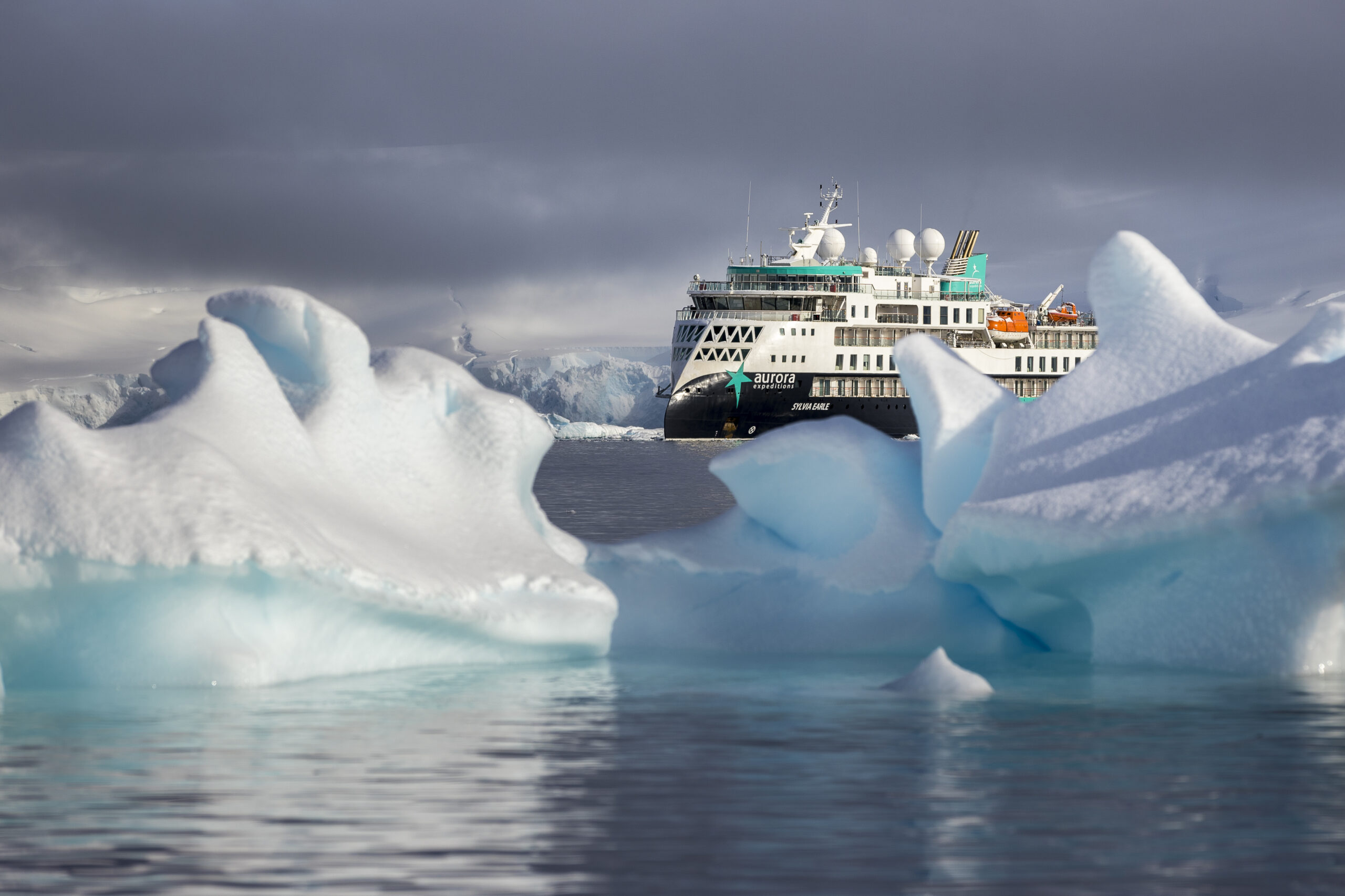 Expedition Cruise Ship Among Arctic Icebergs A white and teal expedition cruise ship navigating through blue-white icebergs in a misty polar seascape with dramatic cloudy skies.