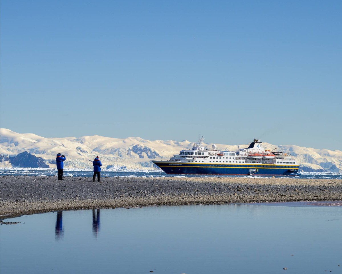 A large expedition cruise ship is anchored in pristine Antarctic waters, with passengers on the rocky beach observing the vessel and snow-covered mountains in the background.