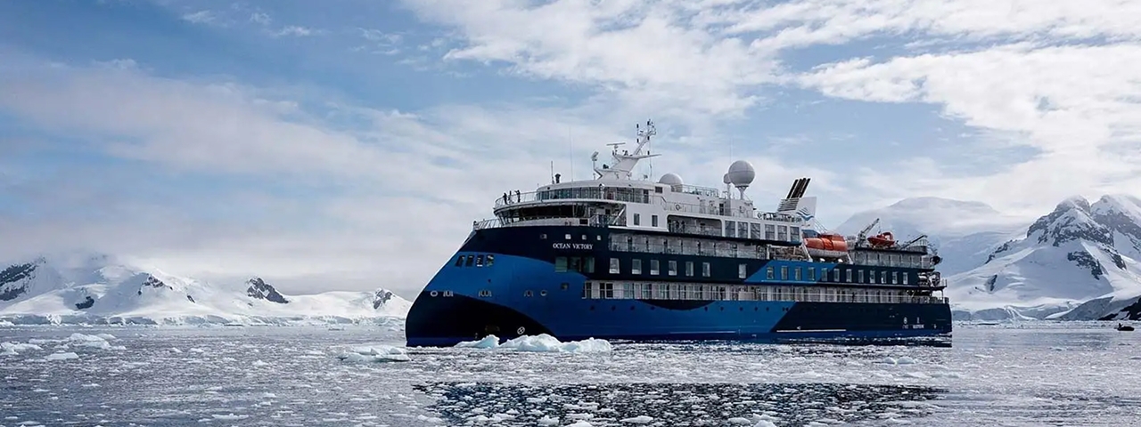 A modern expedition cruise ship with distinctive blue and black hull navigates through icy Antarctic waters surrounded by snow-capped mountains