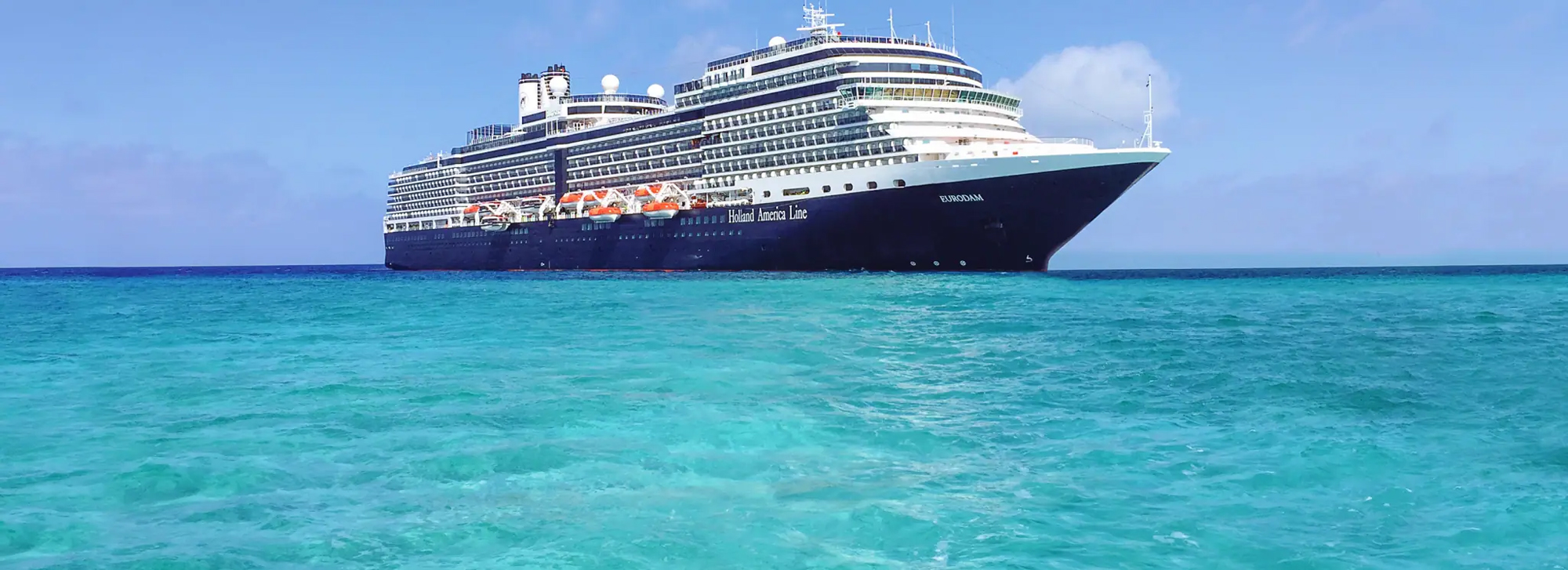 A large blue and white Holland America cruise ship sailing on turquoise Caribbean waters under a clear blue sky.