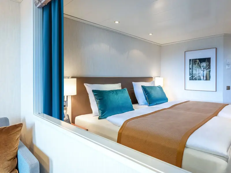 A contemporary cruise ship stateroom featuring a comfortable double bed with teal accent pillows and warm wood furnishings