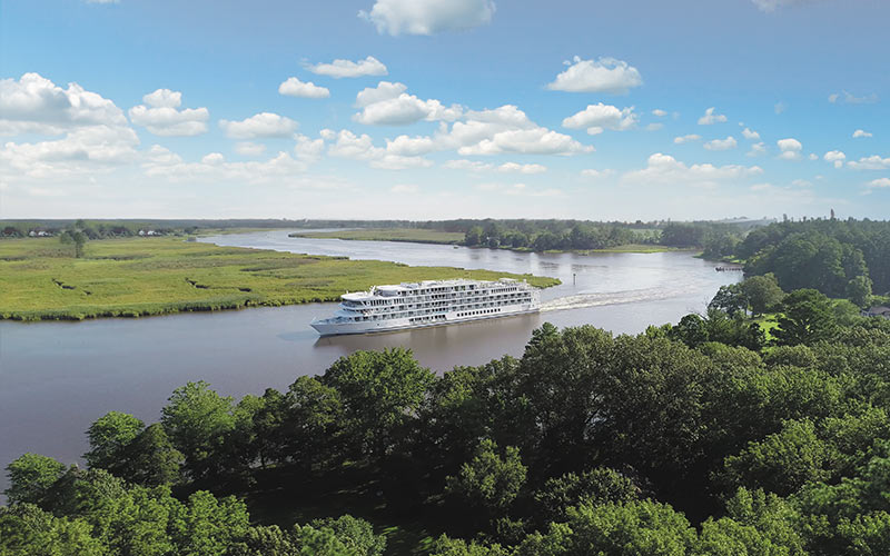 A white river cruise ship navigates through a wide river surrounded by green forests and grassy plains under a blue sky