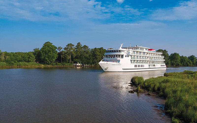 A white river cruise vessel sailing through a serene waterway with lush green trees and grassy shorelines