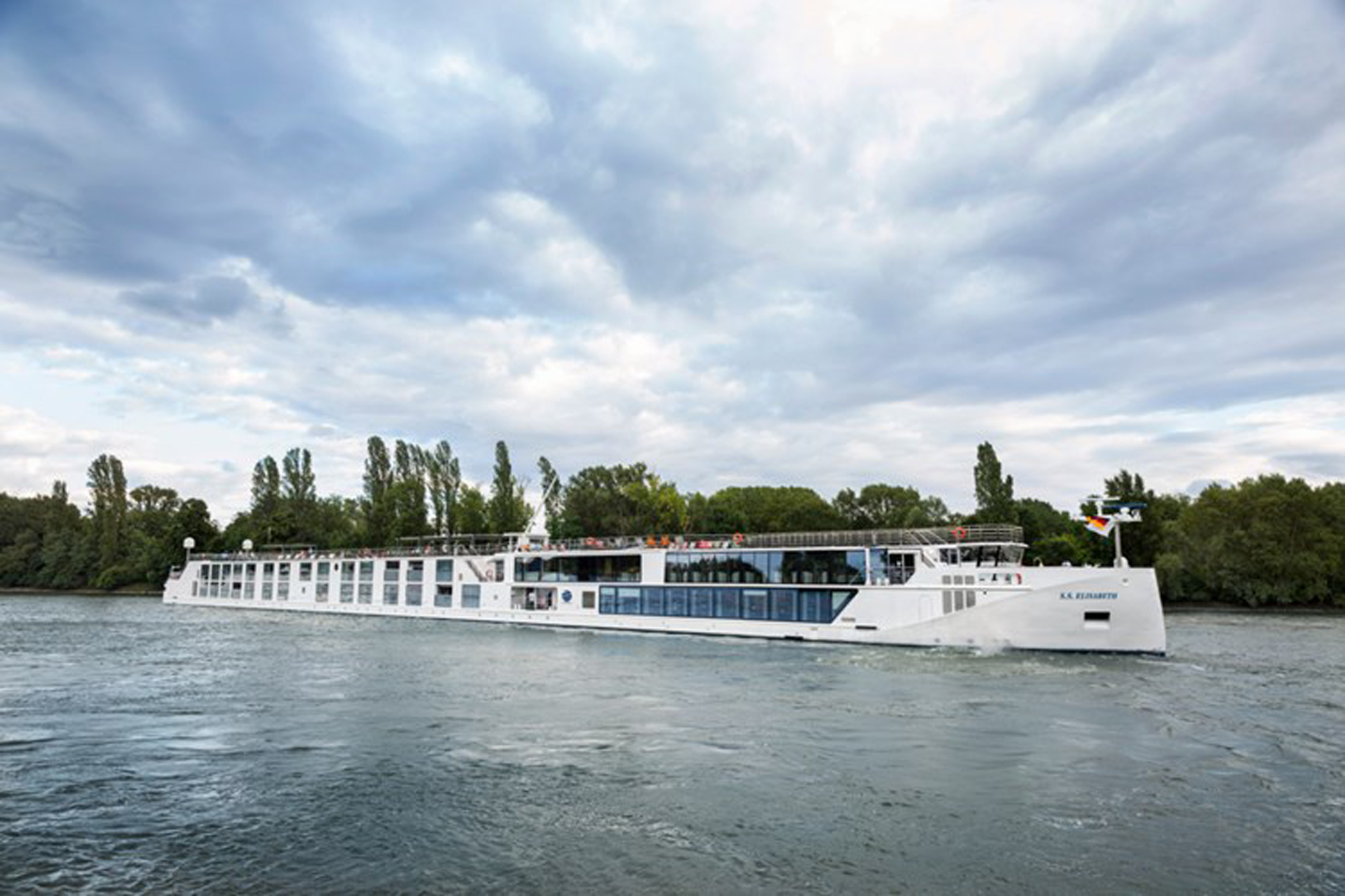 A sleek, white river cruise vessel sailing on a tranquil river, surrounded by lush green trees