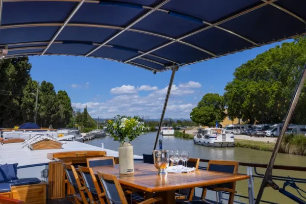 Riverside Dining Deck with Solar Canopy An elegant outdoor dining area situated on a river cruise vessel, featuring a wooden table set for dining under a blue solar panel canopy, with scenic riverside views.