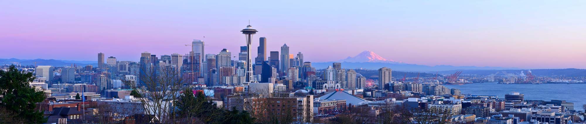 A stunning panoramic view of Seattle's iconic skyline at sunset, featuring the Space Needle as the central landmark, with Mount Rainier visible in the distance and Puget Sound stretching across the foreground. The image captures the city bathed in soft purple and blue twilight hues, showcasing the modern architecture and waterfront setting that define this Pacific Northwest destination.