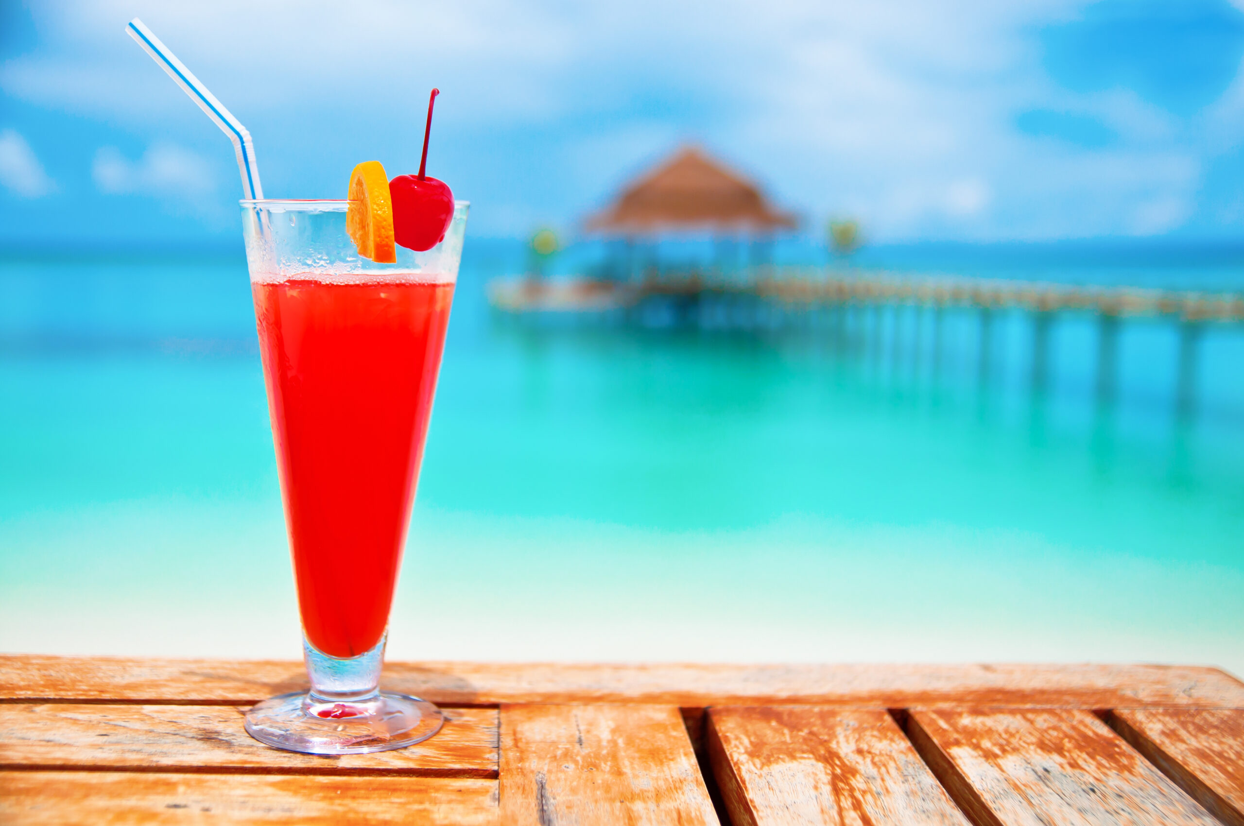 A vibrant red cocktail with ice, garnished with a maraschino cherry and orange slice, sits on a wooden deck overlooking turquoise tropical waters. In the soft-focus background, a thatched-roof beach bungalow and wooden pier are visible against a clear blue sky, evoking a luxurious tropical resort or cruise destination setting.