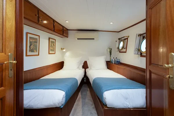 Twin Bed Cabin Aboard Cruise Ship A compact twin-bed cabin aboard a cruise ship featuring two single beds with blue covers, wooden cabinetry, white walls, and nautical-style portholes.