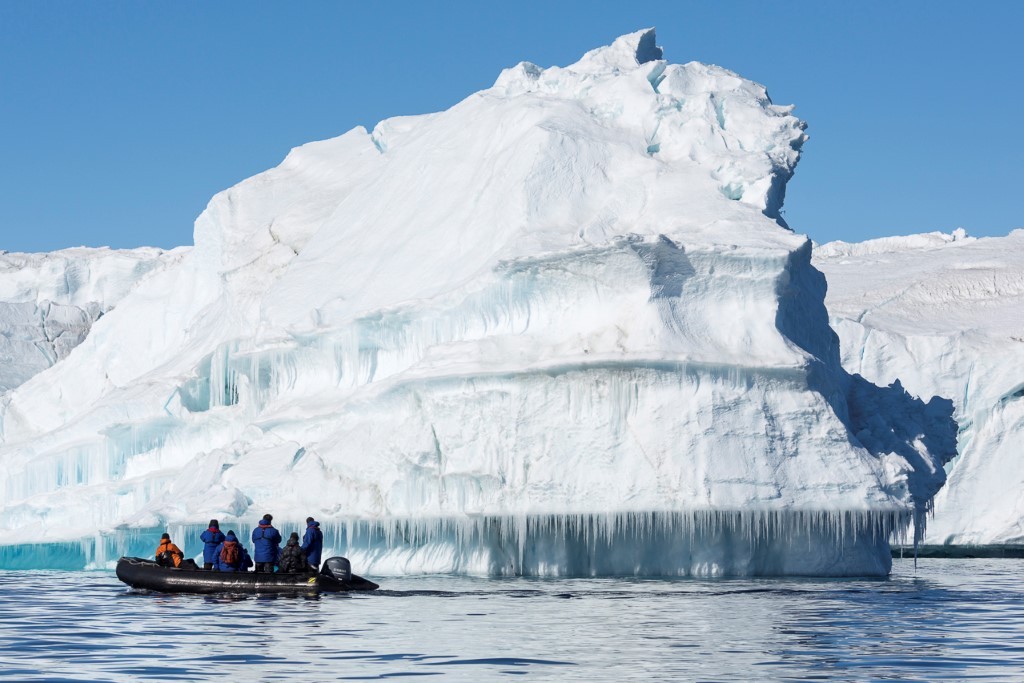 A small zodiac boat carrying adventurers approaches a towering, pristine white iceberg with distinctive blue ice formations and icicle curtains hanging from its base in Arctic waters under clear blue skies.