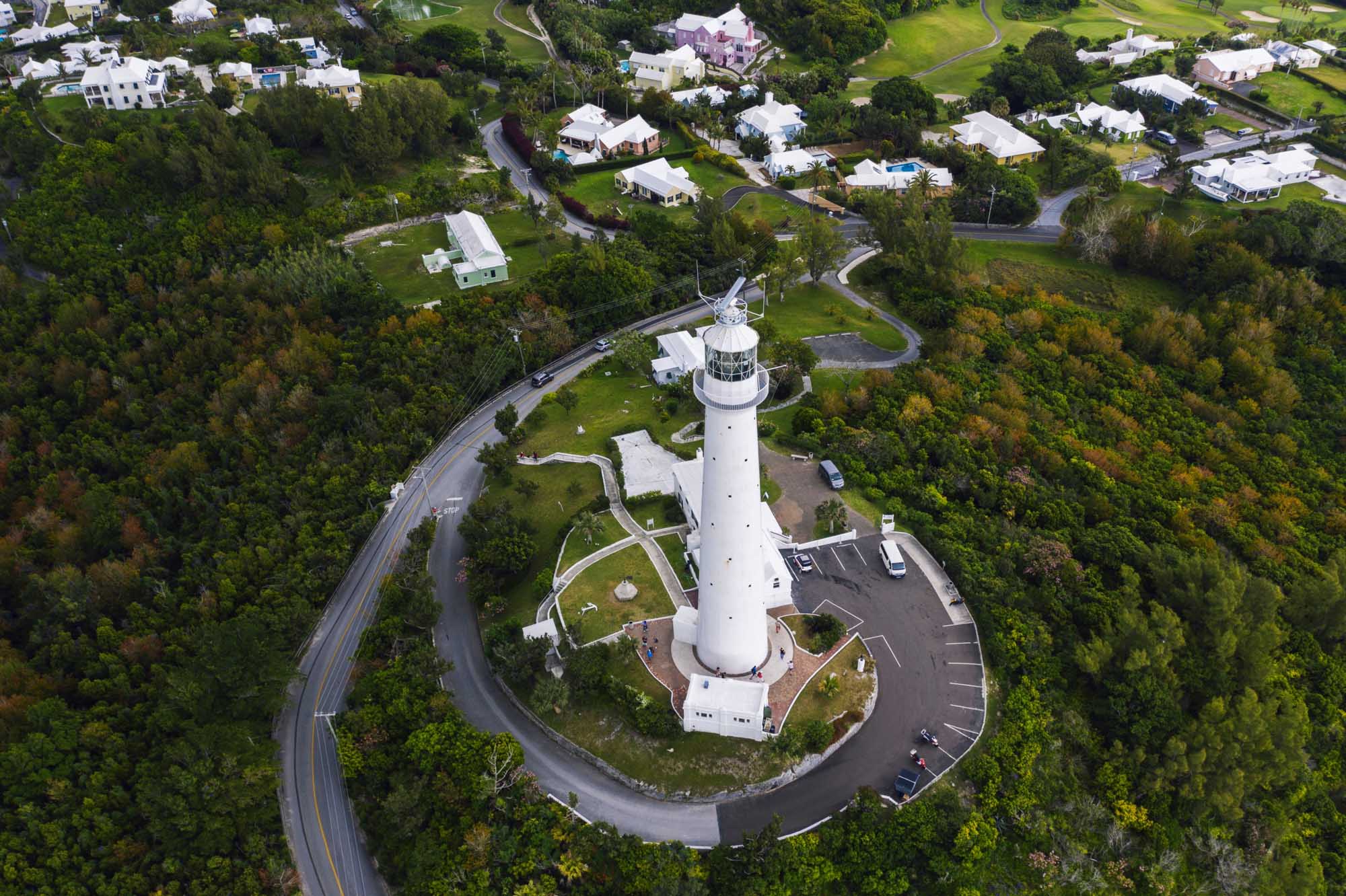 An aerial photograph of a distinctive white lighthouse with its accompanying keeper's residence situated on a promontory in Bermuda, surrounded by lush green vegetation, winding coastal roads, and residential properties.