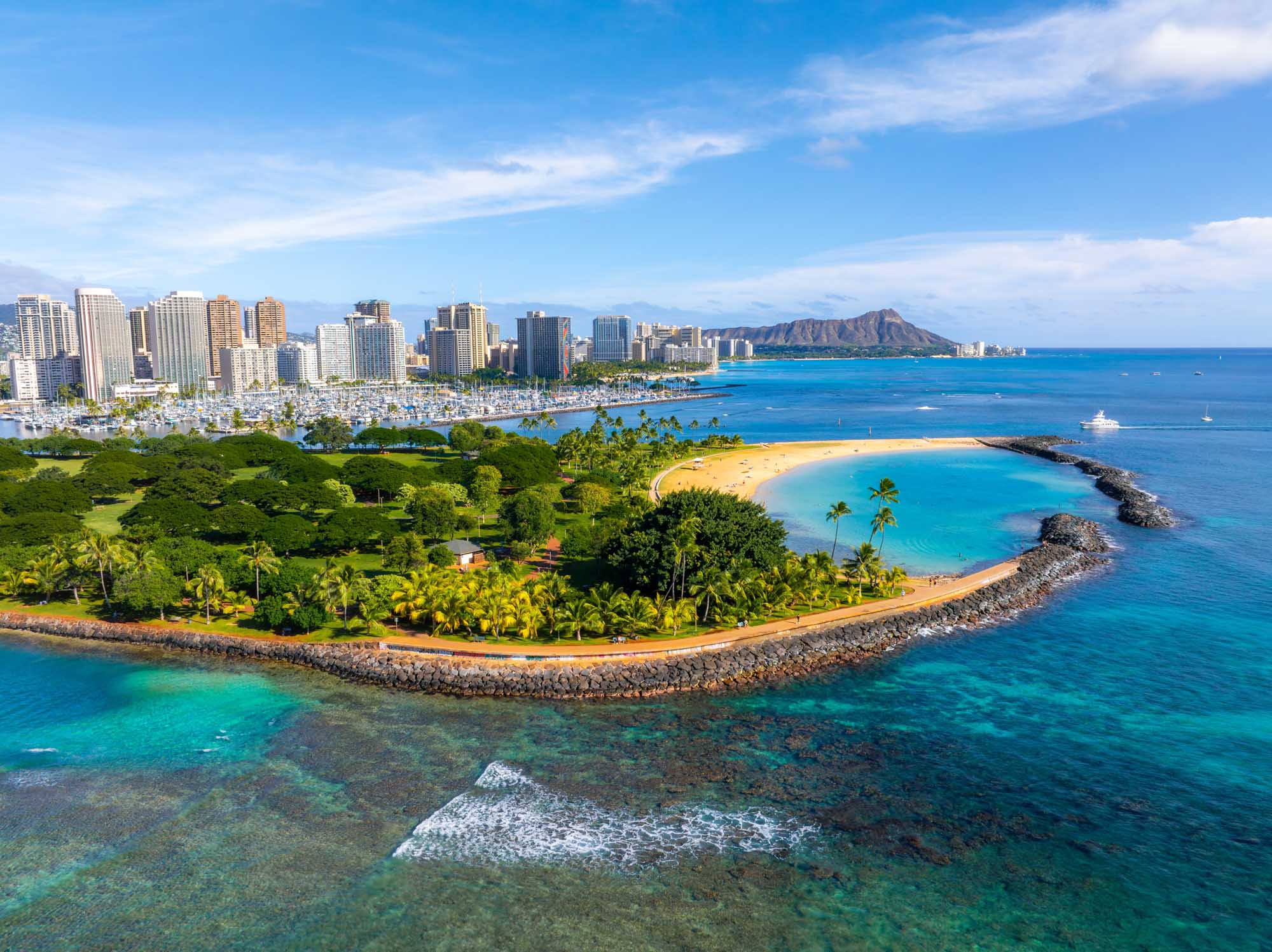 A stunning aerial photograph of Honolulu, Oahu, Hawaii, showcasing the iconic Diamond Head crater, vibrant turquoise waters, sandy beach, lush green park with palm trees, and the modern urban skyline of high-rise buildings. The image captures the perfect blend of natural beauty and urban development that defines Hawaii's capital city.