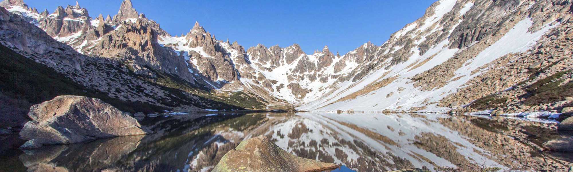 A pristine high-altitude alpine lake mirrors the dramatic jagged peaks and snow-covered slopes of a mountain range, with clear blue sky and scattered patches of snow visible along the rocky terrain.