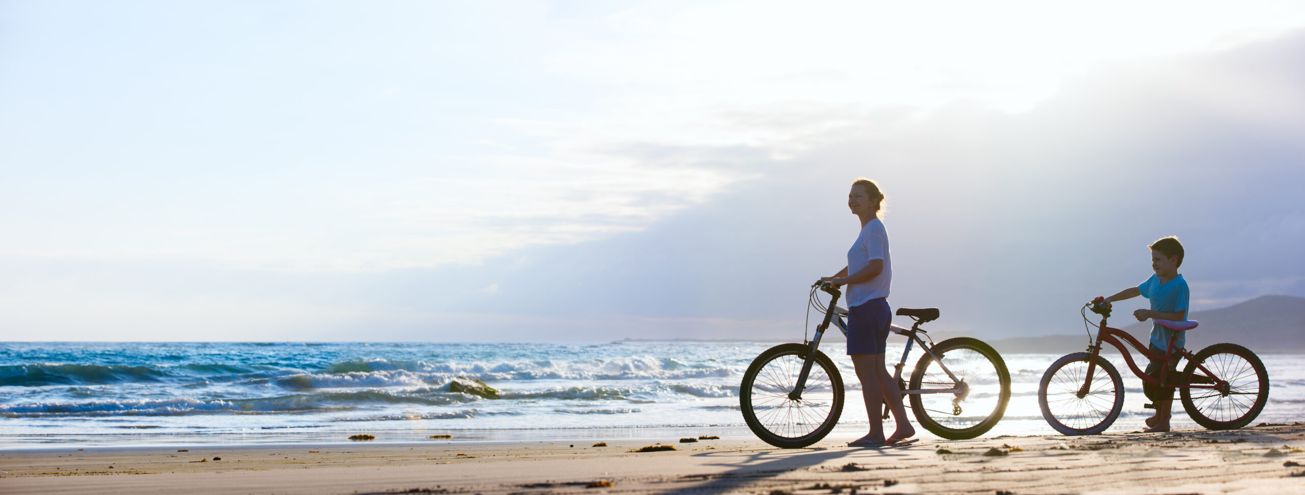 Beachside Cycling Adventure at Sunset An adult and child ride bicycles along a scenic sandy beach with rolling ocean waves in the background during golden hour. The serene coastal landscape features a calm sky with soft clouds, creating an ideal scene for outdoor recreation and family leisure activities.