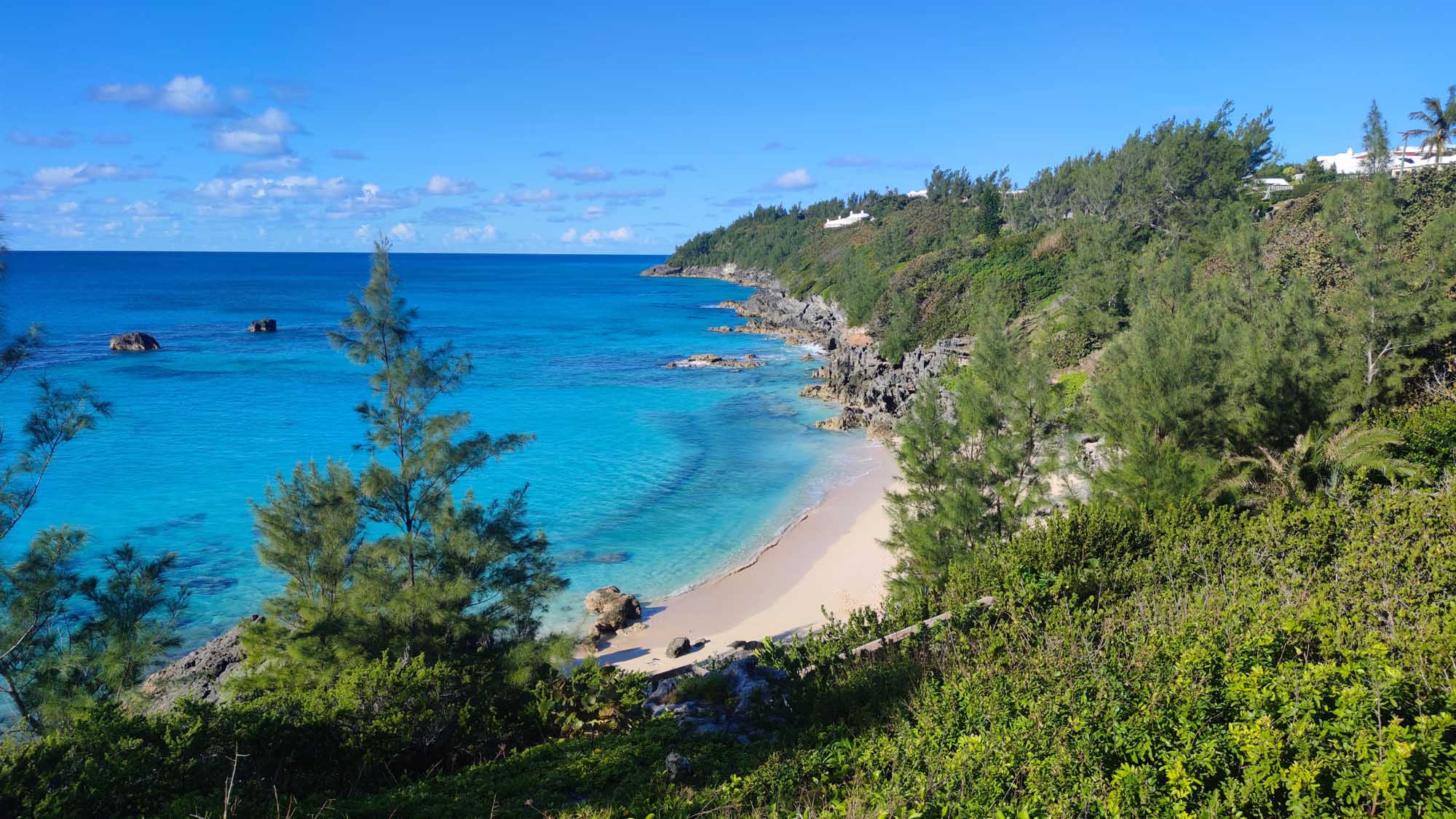 A pristine tropical beach cove nestled between lush green cliffside vegetation and turquoise Atlantic waters, featuring pink-hued sand and rocky outcrops.
