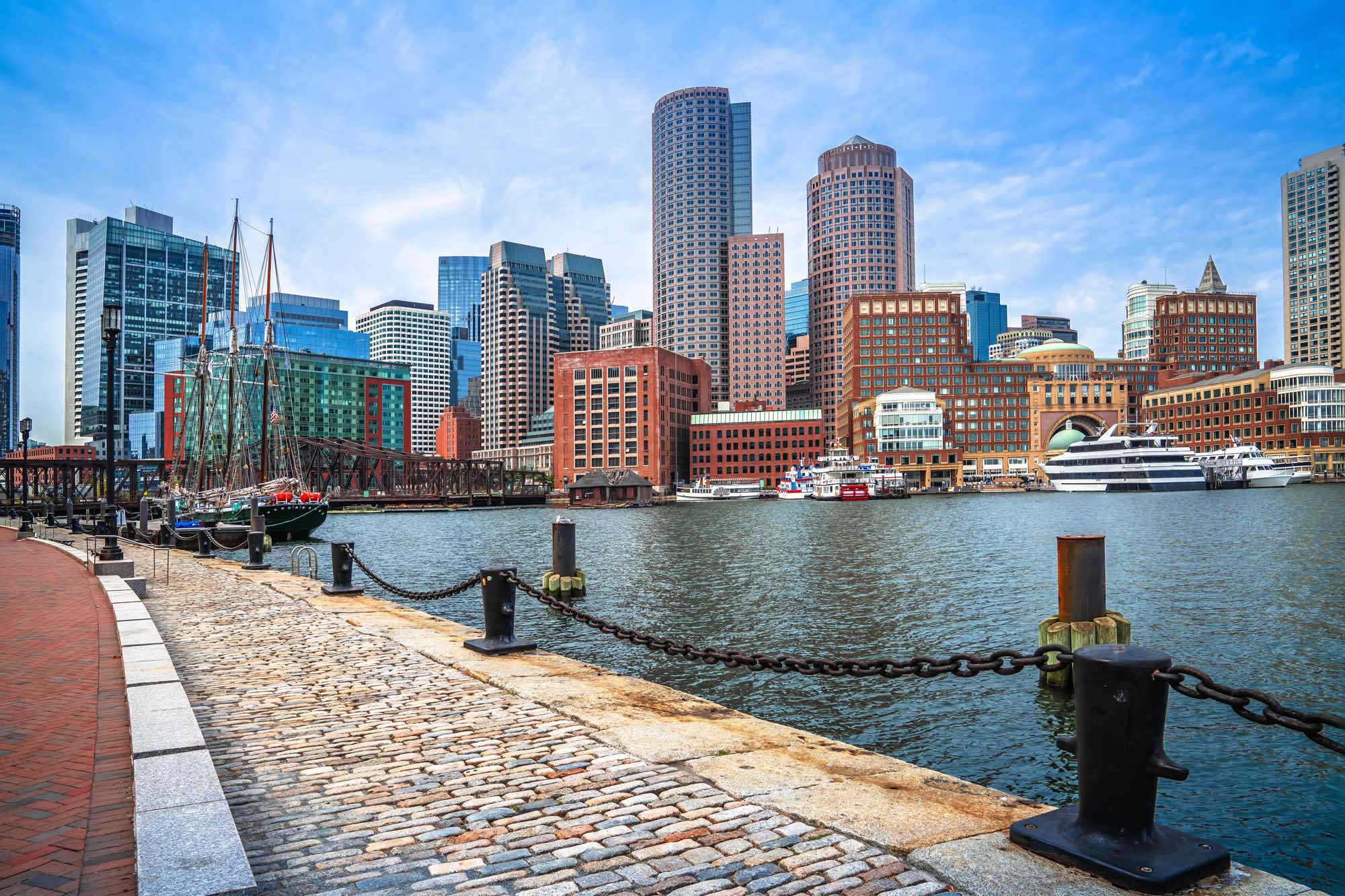 A picturesque view of Boston's waterfront district featuring the iconic cityscape with a mix of historic red-brick buildings and modern glass skyscrapers. The image showcases moored sailing vessels and cruise ships at the pier, with a clear blue sky and calm harbor waters reflecting the vibrant urban landscape.