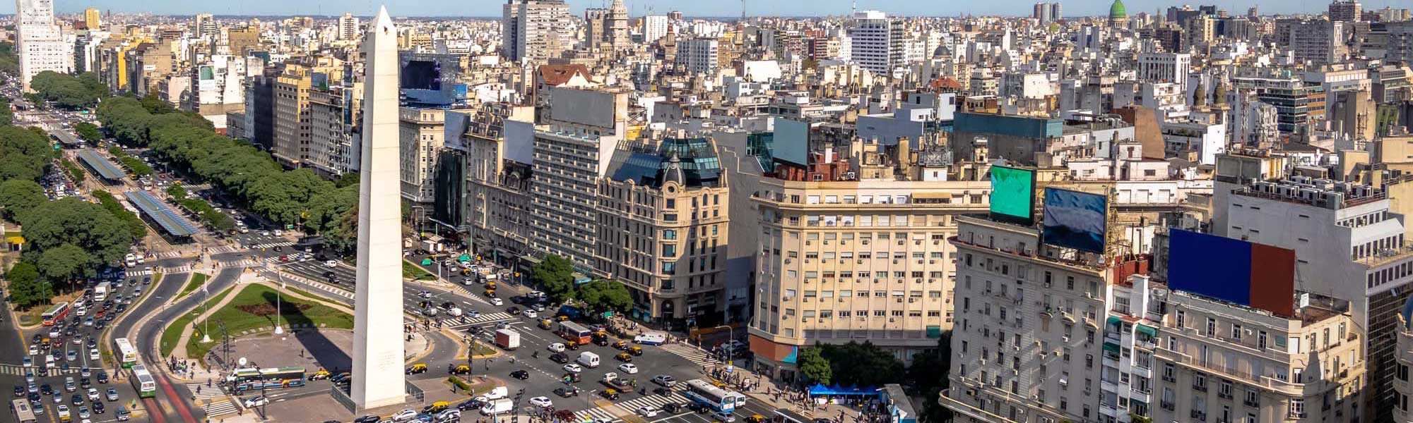 An expansive aerial view of Buenos Aires' iconic downtown district featuring the prominent Obelisco monument at the center of Avenida 9 de Julio. The image showcases the bustling urban landscape with characteristic belle epoque architecture, busy streets filled with vehicles and pedestrians, lush green parks, and the dense cityscape extending to the horizon.