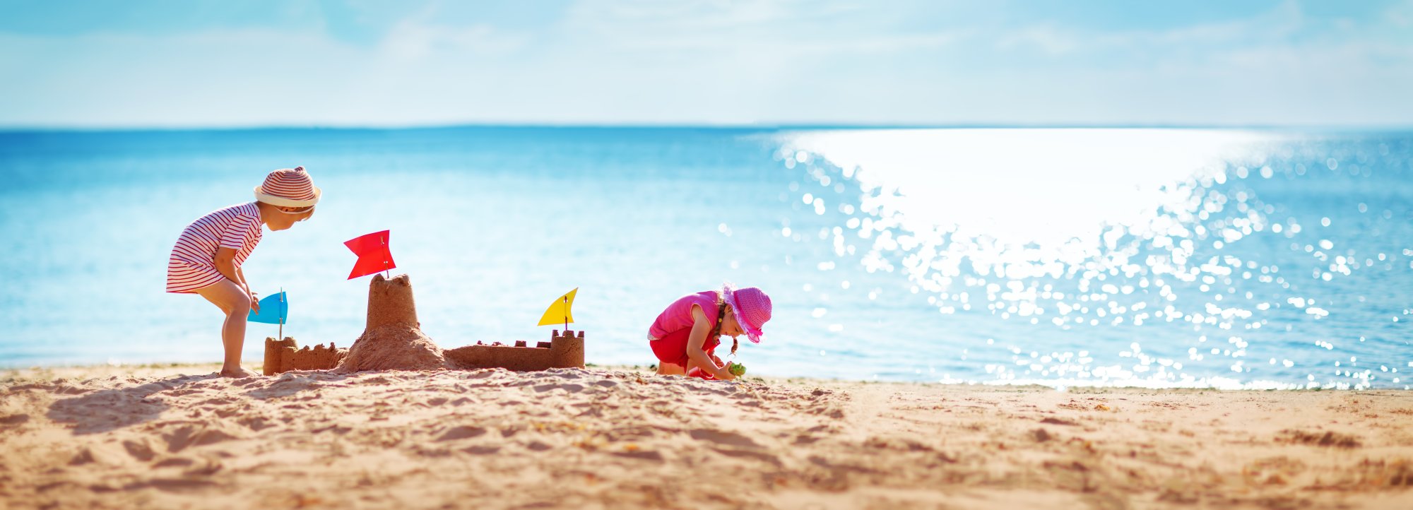 Children Building Sandcastle on Beach Two young children enjoy a sunny beach day, building an elaborate sandcastle with colorful flags while the sparkling turquoise ocean stretches behind them under clear skies.
