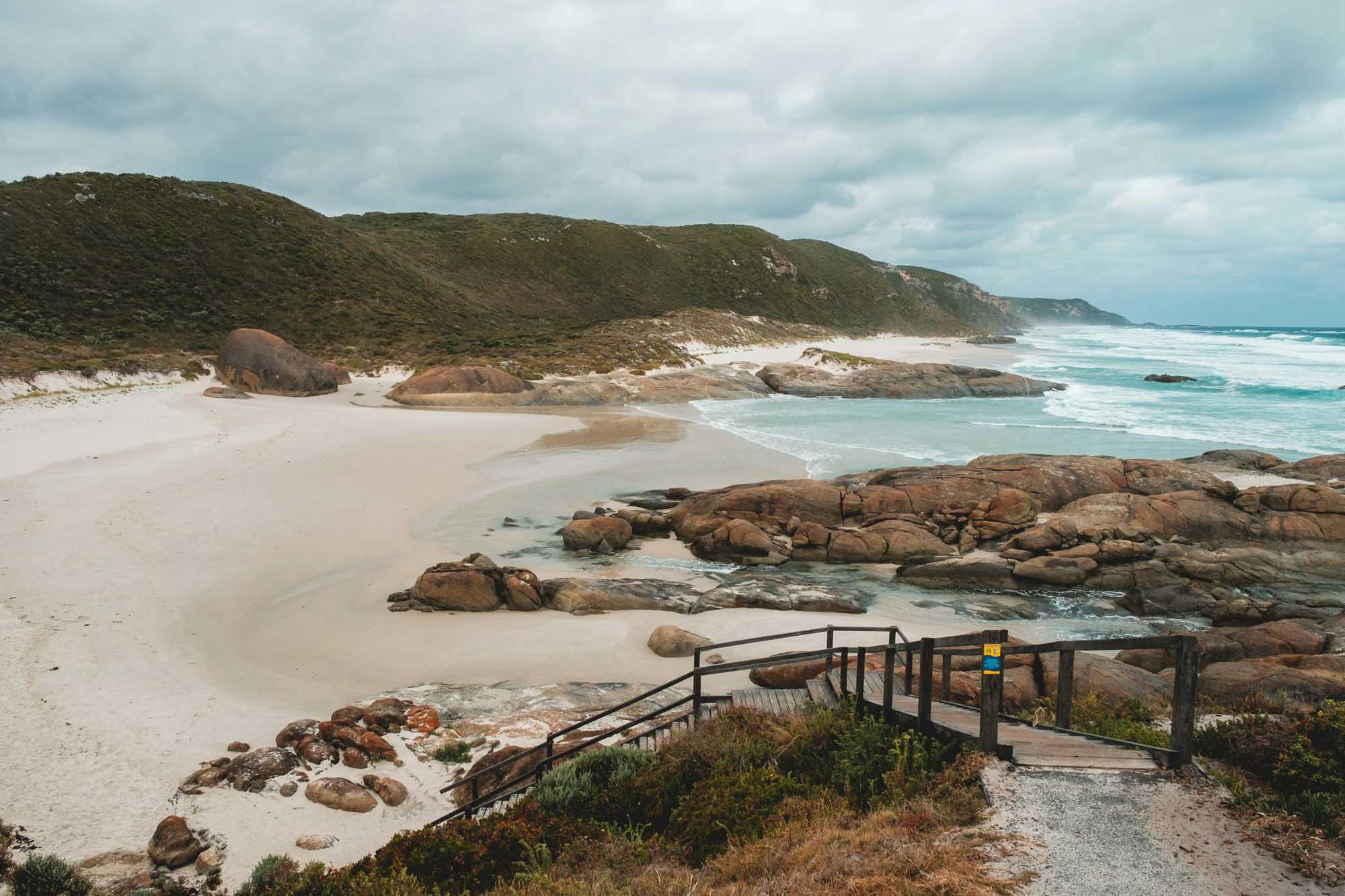 A picturesque coastal landscape featuring a pristine white sand beach framed by distinctive granite rock formations, accessible via a wooden boardwalk. Turquoise waters, lush green hillsides, and dramatic cloud-filled skies complete this scenic seaside destination.
