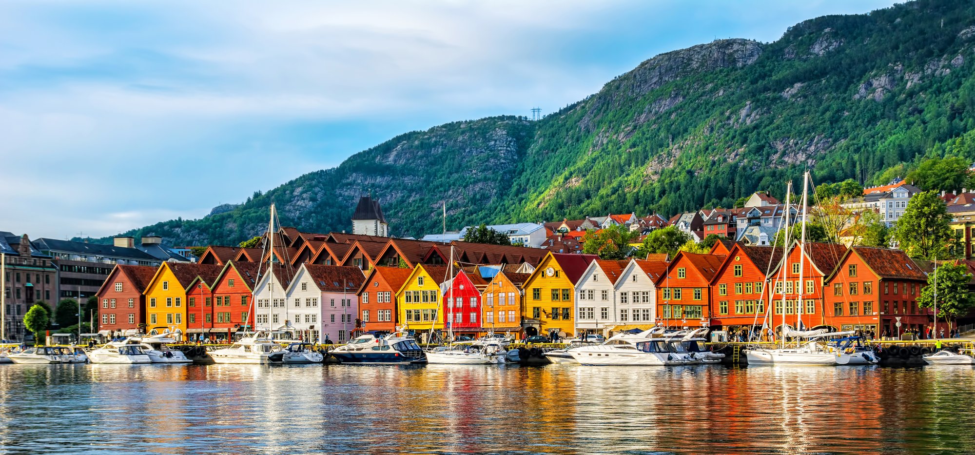 A picturesque Scandinavian waterfront scene featuring a row of vibrant traditional wooden houses in shades of red, orange, yellow, and white, with moored sailboats in the foreground and dramatic forested mountains rising in the background under a clear blue sky.