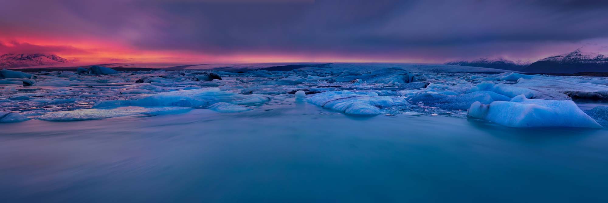 Crimson Afterglow at Jokulsarlon Glacier Lagoon A stunning twilight panorama of Iceland's Jokulsarlon glacier lagoon, featuring icebergs floating in crystalline turquoise water beneath a dramatic sunset sky painted in shades of crimson, purple, and deep blue.