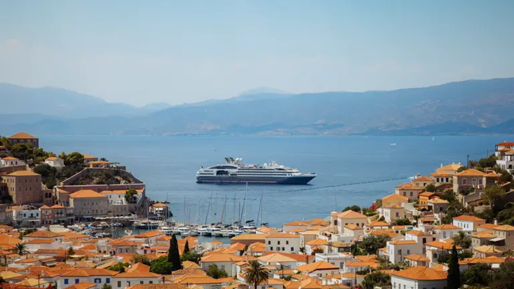 Cruise Ship Anchored at Hydra, Greece A large modern cruise ship anchored in the crystalline Mediterranean waters off the picturesque Greek island of Hydra, with traditional whitewashed buildings featuring orange-tiled roofs cascading down the hillside.