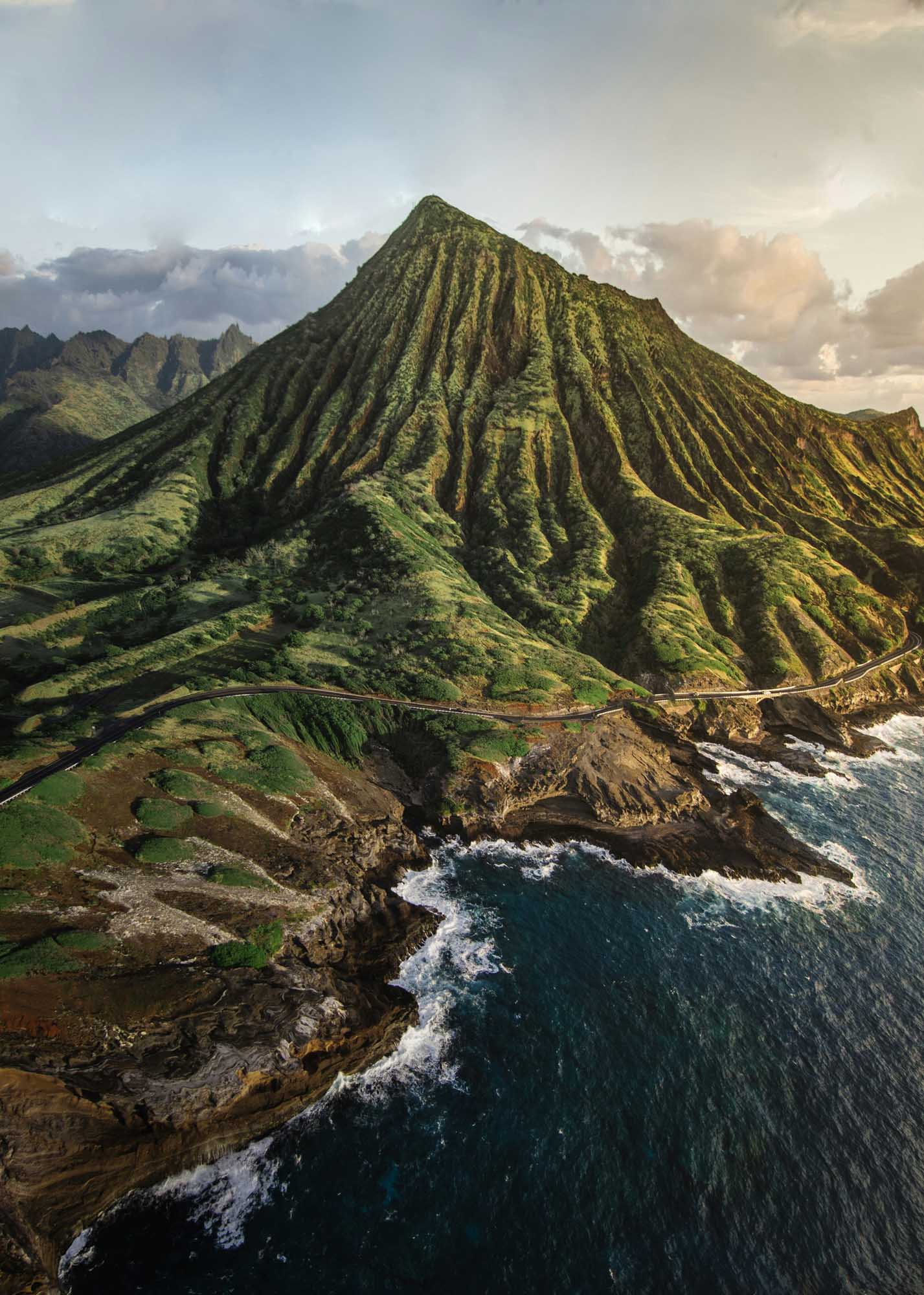 A stunning aerial view of a dramatically fluted volcanic mountain rising from the Hawaiian coastline, with distinctive erosion patterns creating deep vertical ridges across its lush green slopes. Waves crash against the rugged cliffs below while other verdant peaks frame the landscape in the background.