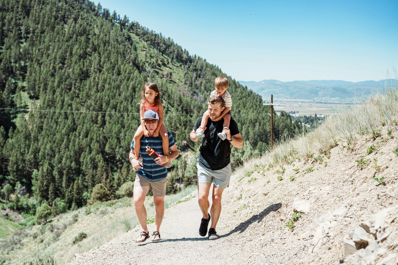 Family Trail Running in Mountain Forest A father carrying his young son on his shoulders runs down a scenic dirt trail through a dense coniferous forest, with two children jogging alongside him. The expansive valley and distant mountains are visible in the background under clear blue skies, capturing an active outdoor family adventure in a mountainous region.