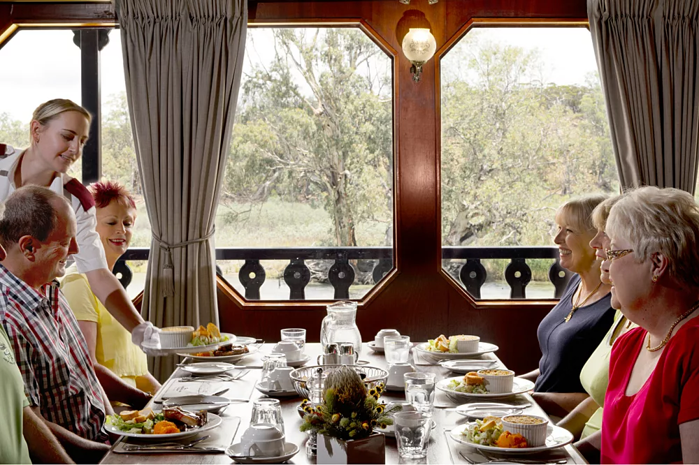 Formal Dining Room with Forest Views An elegant formal dining room aboard a cruise ship with passengers enjoying a meal at a long table. Large windows frame scenic forest and landscape views.