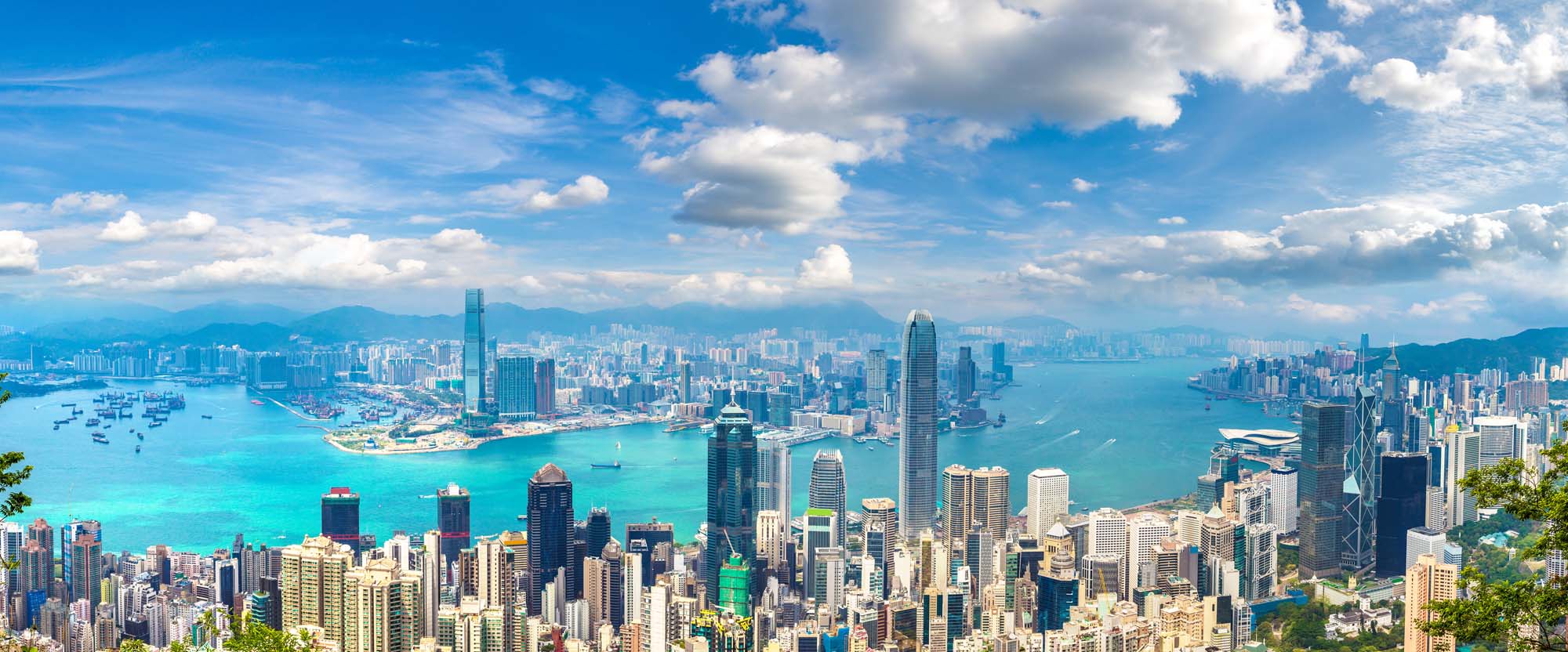 Hong Kong Harbor Panorama with Skyline A stunning panoramic view of Hong Kong's iconic Victoria Harbour and business district skyline, featuring towering skyscrapers, turquoise blue waters dotted with vessels, and lush green mountains in the background under a beautiful blue sky with white clouds.
