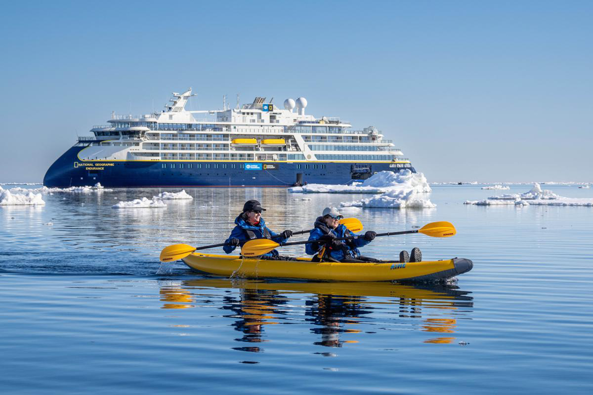 Two kayakers paddle through icy Arctic waters near Svalbard, with the National Geographic Endeavour cruise ship visible in the background surrounded by floating ice floes. The image captures the essence of polar expedition cruising, combining adventure activities with luxury maritime travel.