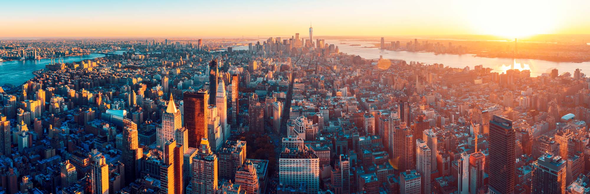 Manhattan Aerial Panorama at Sunset An expansive aerial panoramic view of Manhattan captured during golden hour, showcasing the iconic skyline with illuminated skyscrapers, the East River, Hudson River, and surrounding boroughs bathed in warm sunset light.