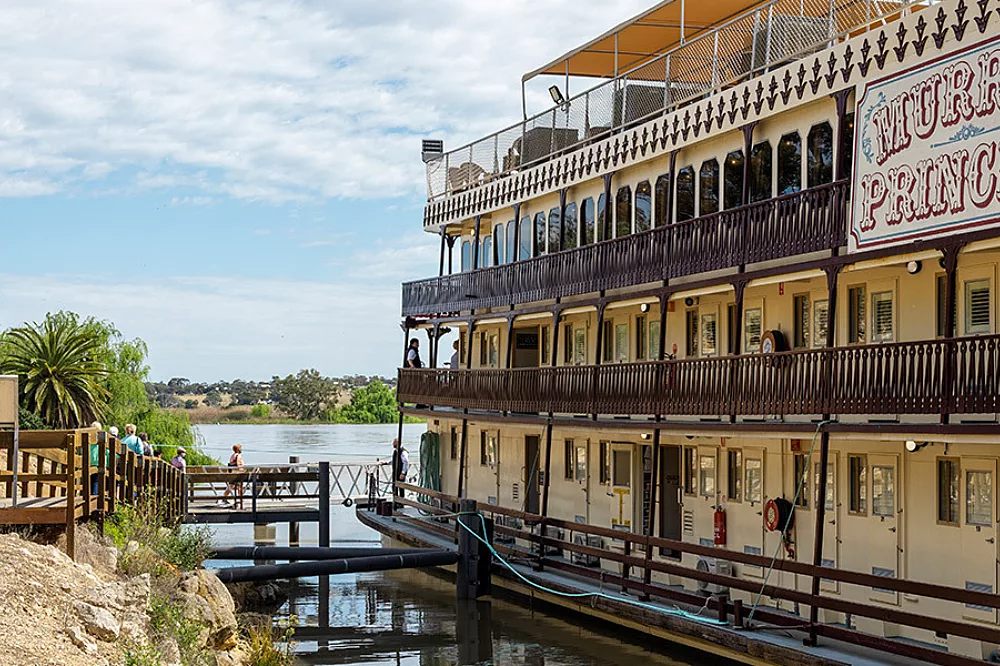 Murray Princess Riverboat Docked at Riverbank A three-deck paddlewheel riverboat with cream and brown colonial-style architecture docked along a peaceful river, featuring ornate railings and multiple levels of verandahs.