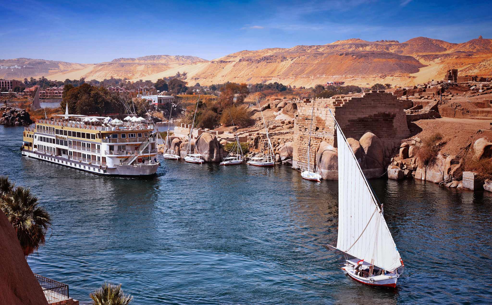 Nile River Cruise Ship at Aswan A large river cruise ship docked along the Nile River at Aswan, Egypt, with traditional felucca sailboats and ancient stone ruins visible along the riverbank.