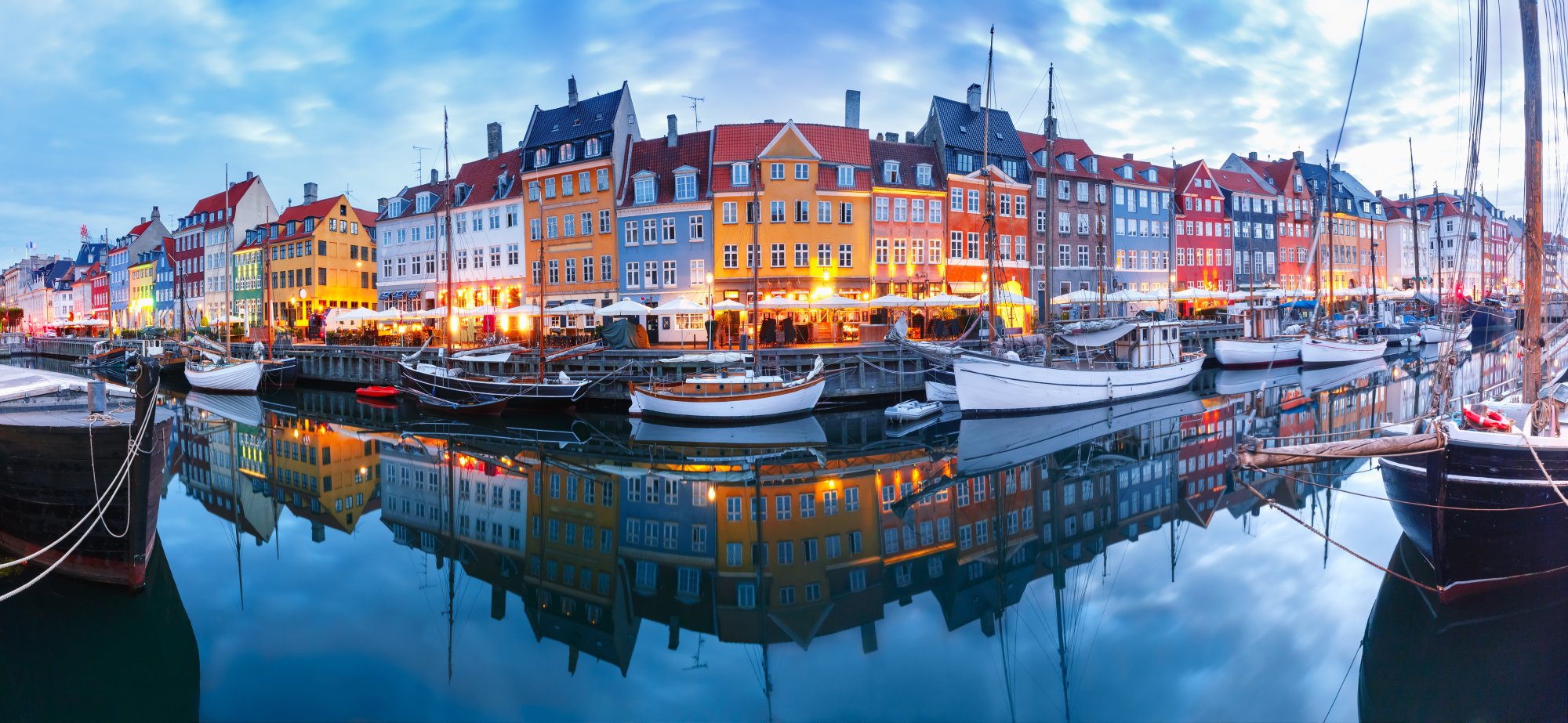 Nyhavn Harbor at Dusk with Colorful Buildings A stunning twilight view of Copenhagen's iconic Nyhavn waterfront, featuring a row of vibrant multi-colored 17th-century buildings reflected perfectly in the still harbor waters, with traditional sailing boats moored alongside the quay and warm evening lights illuminating the historic facades.