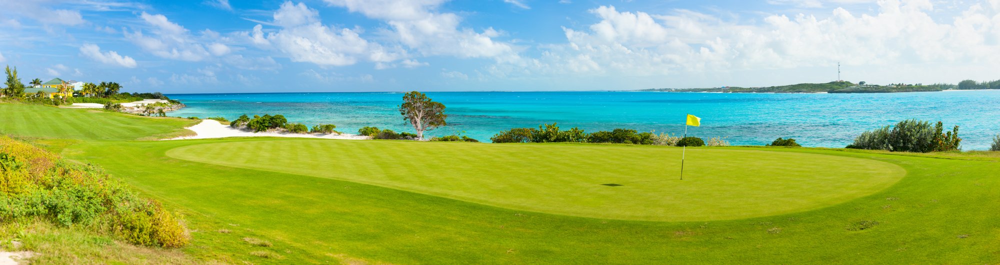 A scenic coastal golf course featuring a pristine green fairway overlooking crystal-clear turquoise waters and a white sand beach, with lush tropical vegetation.