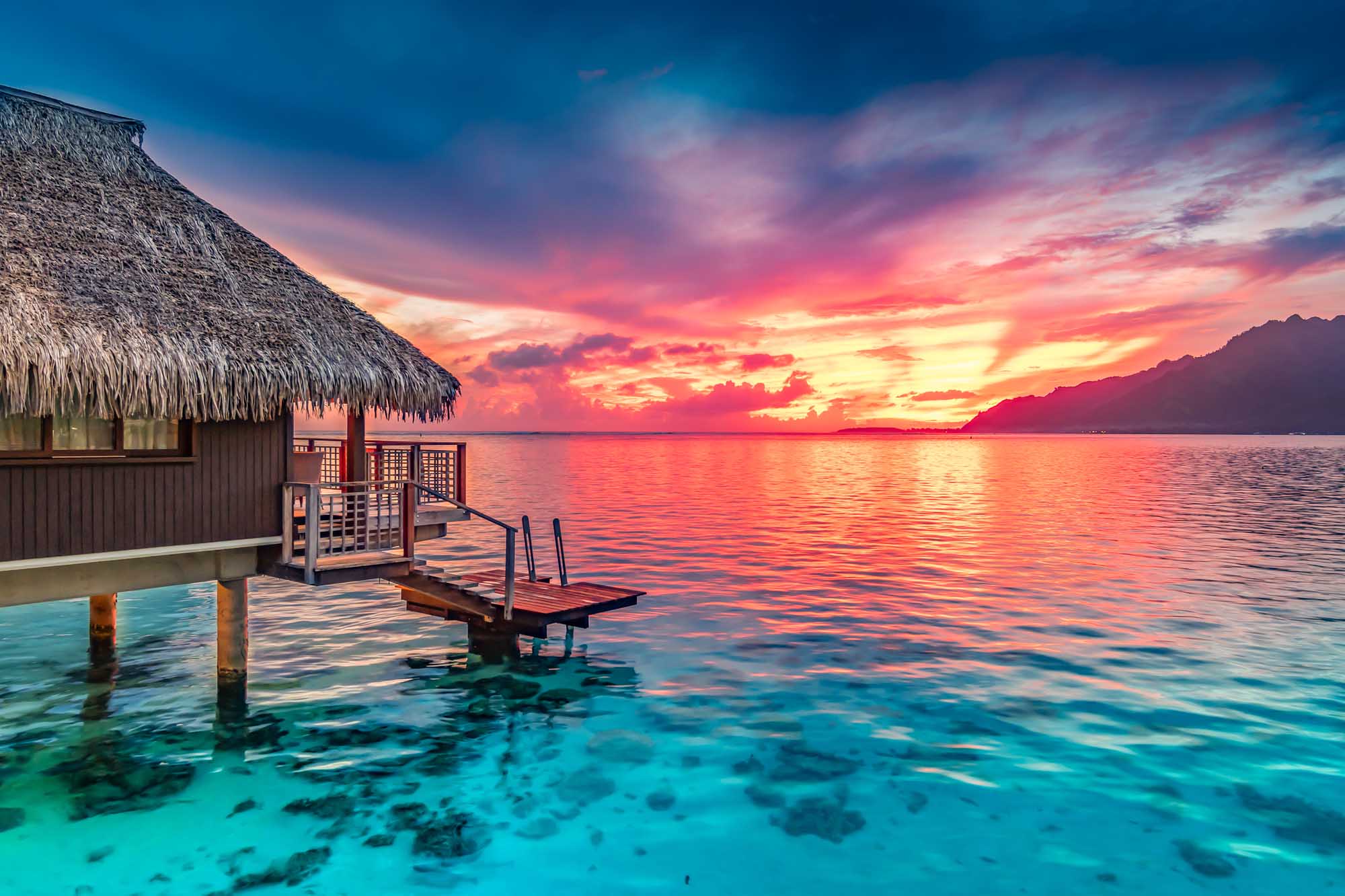 Overwater Bungalow Sunset in Moorea A stunning tropical sunset scene featuring a traditional thatched-roof overwater bungalow with wooden deck and jetty in crystal-clear turquoise waters. The dramatic sky displays vibrant hues of pink, orange, and gold reflecting across the calm ocean, with distant volcanic mountains silhouetted on the horizon.